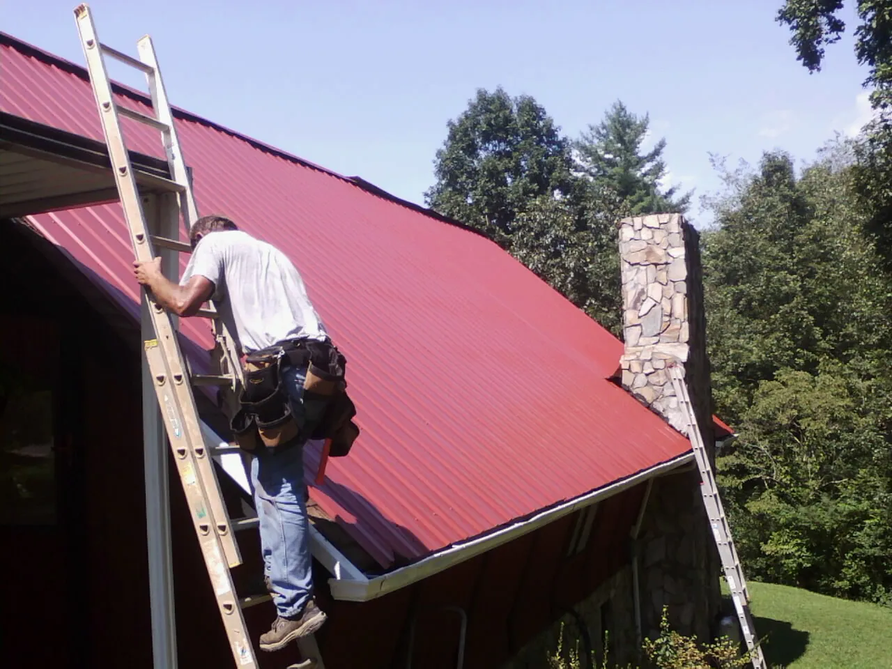 Roofer on a ladder beside a red metal roof and stone chimney — Soffit & Fascia Repair by Roofing Brothers in Wichita